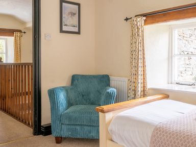 A bedroom corner with a blue armchair next to a bed and a window with floral curtains at Hilltop in Graythwaite near Hawkshead