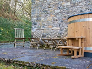 An outdoor stone patio with wooden chairs and a wooden hot tub next to a stone wall at Hilltop in Graythwaite near Hawkshead