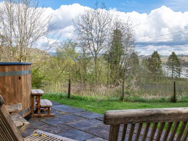 A patio with wooden chairs and a hot tub overlooking trees and distant hills at Hilltop Graythwaite near Hawkshead