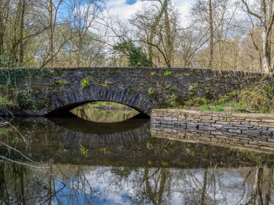 A stone arch bridge over a calm river with trees and vegetation around at Hilltop in Graythwaite near Hawkshead