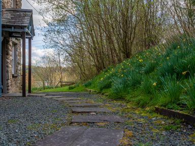 A stone path leading past a stone house with a garden of green plants and yellow flowers at Hilltop in Graythwaite near Hawkshead