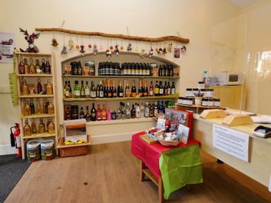 A shop corner with shelves of bottles and jars a small table with products and a counter with a protective screen at Mill Cottage in Graythwaite near Hawkshead