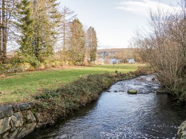A river flowing beside grassy land with trees and a lake in the background at Mill Cottage in Graythwaite near Hawkshead