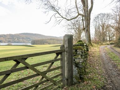 A wooden gate leading to a grassy field with a dirt path and stone wall beside trees at Mill Cottage in Graythwaite near Hawkshead