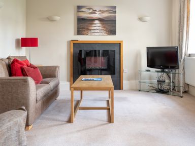 A living room with a sofa red pillows a wooden coffee table fireplace and a television on a glass stand at Mill Cottage in Graythwaite near Hawkshead