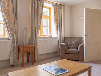 A room with two windows with curtains a wooden side table with a vase a brown armchair an open door and a wooden coffee table with a magazine Mill Cottage Graythwaite near Hawkshead
