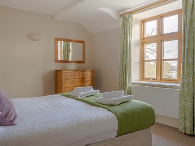 A bedroom with a bed, green curtains, a wooden chest of drawers, and a window at Mill Cottage in Graythwaite near Hawkshead