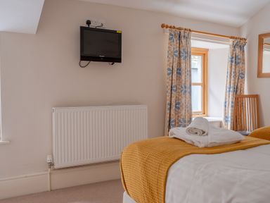 A bedroom with a wall-mounted TV radiator a window with floral curtains a wooden chair and a bed with folded towels at Mill Cottage Graythwaite near Hawkshead