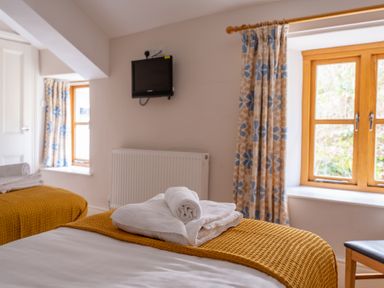 A bedroom with two beds covered with mustard blankets and white towels on them a small wall-mounted television and windows with floral curtains at Mill Cottage in Graythwaite near Hawkshead