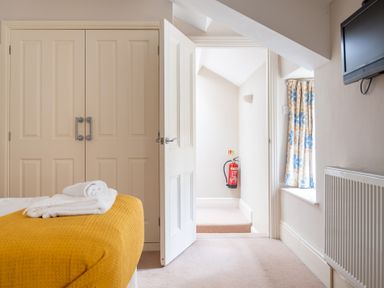 A bedroom with a bed covered by a yellow blanket folded towels a closet door a partially open door a fire extinguisher on a wall and a window with floral curtains at Mill Cottage in Graythwaite near Hawkshead