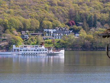 A passenger boat named Teal on a lake with tree-covered hills and buildings in the background at Hammerhole in Graythwaite near Hawkshead