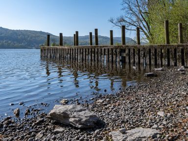 A lakeshore with a wooden pier extending into the water and hills in the background at Hammerhole in Graythwaite near Hawkshead