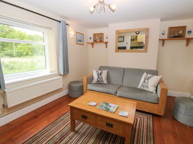 A living room with a grey sofa, wooden coffee table, striped rug, window with curtains, and wall shelves at 2 Lock Cottages in Honing near East Ruston