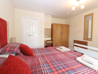 A bedroom with a double bed with red and plaid bedding a wooden wardrobe a small desk with a chair a wall mirror and folded towels on the bed at 2 Lock Cottages Honing near East Ruston