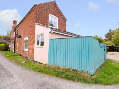 A brick house with a blue wooden fence and a small patch of grass near a road at 2 Lock Cottages in Honing near East Ruston