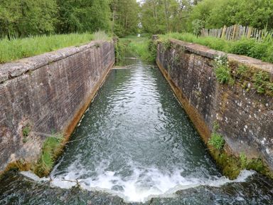 A narrow canal lock with brick walls and flowing water surrounded by trees and greenery at 2 Lock Cottages in Honing near East Ruston