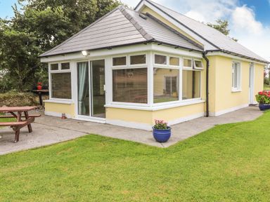 A yellow house with large windows and a concrete path with grass and potted plants at Moybella Lodge in Lisselton near Ballybunion County Kerry