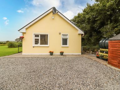 A yellow single-story house with two windows a gravel driveway and flower pots at Moybella Lodge in Lisselton near Ballybunion County Kerry