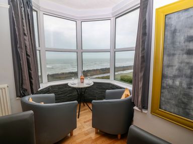 A corner seating area with two grey chairs and a small round table with glasses and a bottle by a large window overlooking the sea at Morolwg in Llanaber near Barmouth