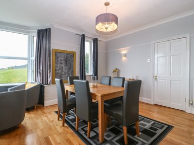 A dining room with a wooden table and six chairs and two armchairs near windows at Morolwg in Llanaber near Barmouth