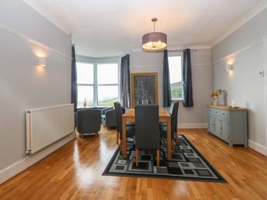 A dining room with a wooden floor a dining table with chairs a sideboard curtain covered windows and a seating area by a bay window at Morolwg in Llanaber near Barmouth