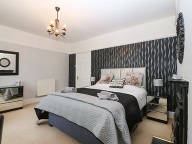 A bedroom with a double bed two bedside tables with lamps a chandelier a dresser with a mirror and patterned wallpaper at Morolwg in Llanaber near Barmouth