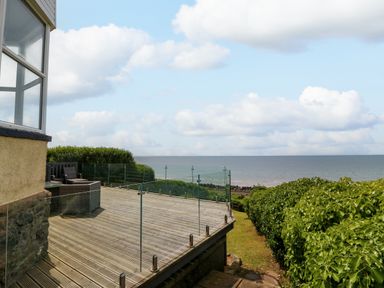 A wooden deck with glass railing overlooking the sea and a green hedge at Morolwg in Llanaber near Barmouth