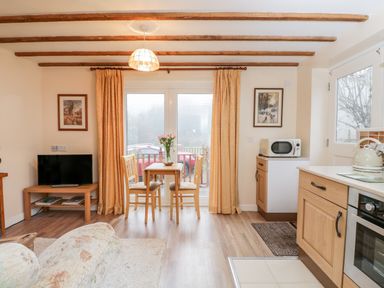 A dining area with a small table and two chairs next to sliding glass doors with curtains a kitchen counter with a microwave and kettle and a television on a wooden stand at Woods Edge in Mitcheldean