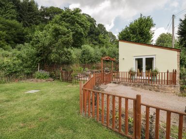 A small fenced garden area with a gravel path next to a yellow building surrounded by trees and greenery at Woods Edge in Mitcheldean