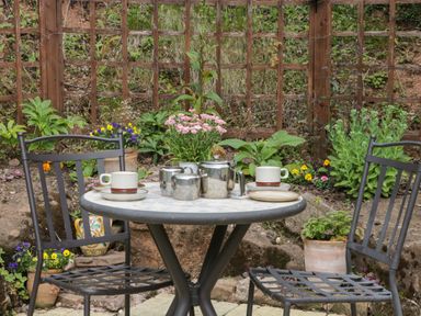 An outdoor patio table with tea set and two metal chairs surrounded by plants and flowers at Woods Edge in Mitcheldean