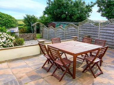 An outdoor patio with a wooden table and six chairs next to a garden with plants and a wooden fence at The Grange in Little Haven near Haverfordwest