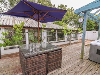 An outdoor wooden deck with a wicker table and purple umbrella next to a white building with windows and a blue pergola at Cariad Cottage in Cross Inn