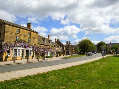 A street with stone buildings covered in purple flowers cars on the road and grass area with trees at Pennylands Willow Lodge in Broadway