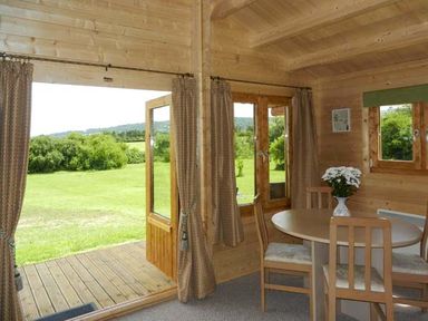 A wooden dining area with a round table and four chairs near open glass doors leading to a grassy outdoor area at Pennylands Willow Lodge in Broadway