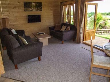 A living room with two dark sofas a coffee table a wall-mounted TV and open double doors leading outside at Pennylands Willow Lodge in Broadway