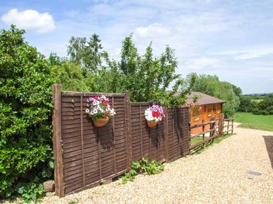 A gravel pathway with wooden fence and hanging flower baskets next to a wooden building in a rural setting at Pennylands Willow Lodge in Broadway