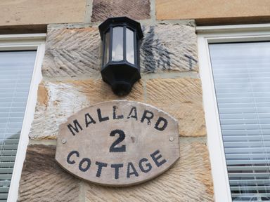 A wooden sign with the text Mallard 2 Cottage mounted on a stone wall under a black outdoor light fixture at Mallard Cottage in Whitby