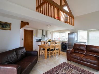 An open plan living area with two brown leather sofas a wooden dining table with chairs kitchen appliances and a balcony railing above at Mallard Cottage in Whitby