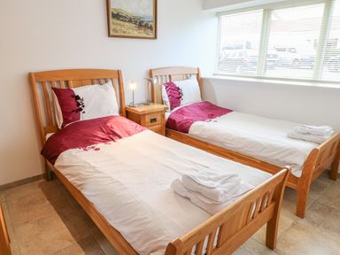 A bedroom with two single wooden beds with white and red bedding a nightstand with two lamps and a window with blinds at Mallard Cottage in Whitby