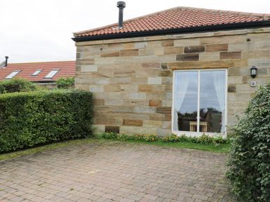 A stone building with a tiled roof and a sliding glass door with curtains in front of a paved area at Mallard Cottage in Whitby