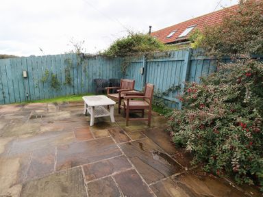 An outdoor patio with stone tiles two wooden chairs a white table blue wooden fencing and flowering bushes at Mallard Cottage in Whitby