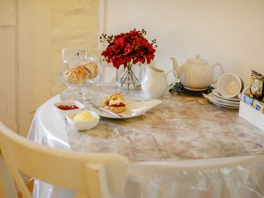 A kitchen table set with scones jam and cream a teapot cups a milk jug and a vase of red flowers at Wagtail Cottage in East Taphouse