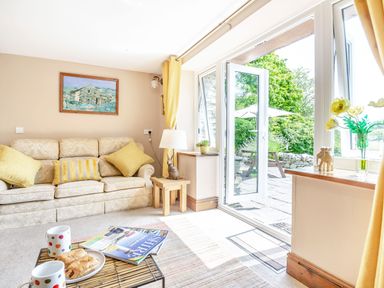 A living room with a sofa yellow cushions a small wooden table with a lamp an open door to a garden and a coffee table with pastries and mugs at Bwythyn Y Wennol in Nantmor near Beddgelert