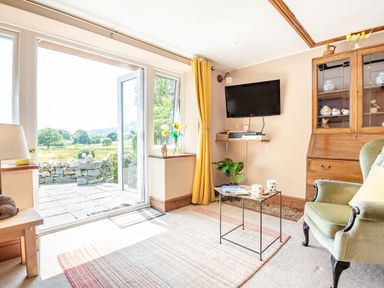 A living room with an open glass door leading outside a wooden cabinet a green armchair and a wall-mounted television at Bwythyn Y Wennol in Nantmor near Beddgelert 