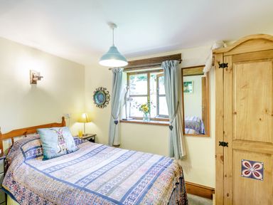 A bedroom with a bed covered with a patterned quilt beside a wooden wardrobe and a window with curtains at Bwythyn Y Wennol in Nantmor near Beddgelert