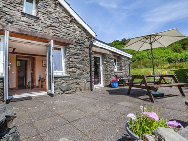 Stone house exterior with open doors leading inside a covered patio with table bench and umbrella at Bwythyn Y Wennol in Nantmor near Beddgelert