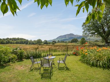 A garden with a round table and four chairs surrounded by grass and flowers with a mountain in the background at Bwythyn Y Wennol in Nantmor near Beddgelert