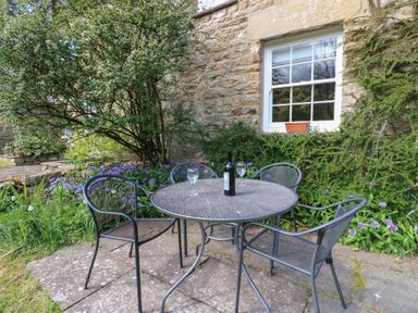 An outdoor patio area with a round metal table and four metal chairs with a bottle and two glasses on the table next to a stone building with a window and surrounding greenery at Farrier's Cottage in Garsdale