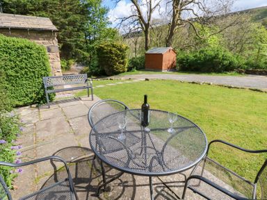 A garden patio with a metal table and chairs a wine bottle and glasses on the table a wooden shed and greenery in the background at Farrier's Cottage in Garsdale