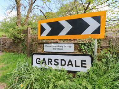 A road sign with arrows a please drive slowly sign and a garsdale village sign surrounded by grass and trees